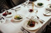 A table set with a white table cloth and containing glasses of red wine and luxurious dishes like rack of lamb and filet mignon