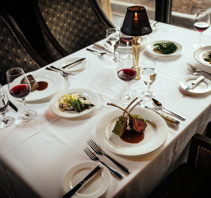 A table set with a white table cloth and containing glasses of red wine and luxurious dishes like rack of lamb and filet mignon